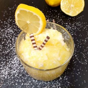 Gelbe Zitronencreme mit untergehobenem Eischnee in eines Dessertglas, dekoriert mit einer Zitronenscheibe und Schkoladenstäbchen